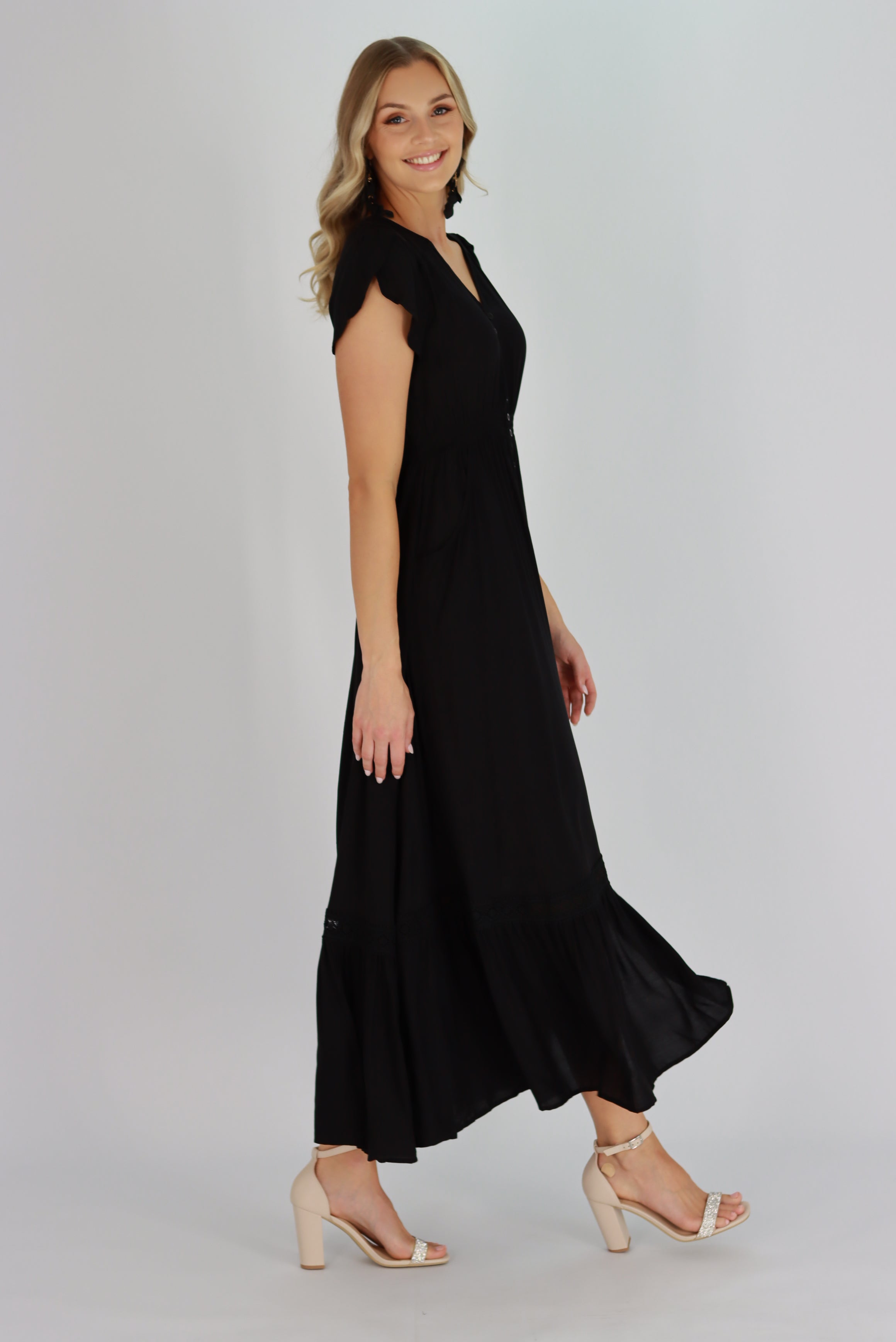 A woman wearing a black maxi dress with a V-neckline, short sleeves, and front functional buttons. The dress has a front split and two side pockets.