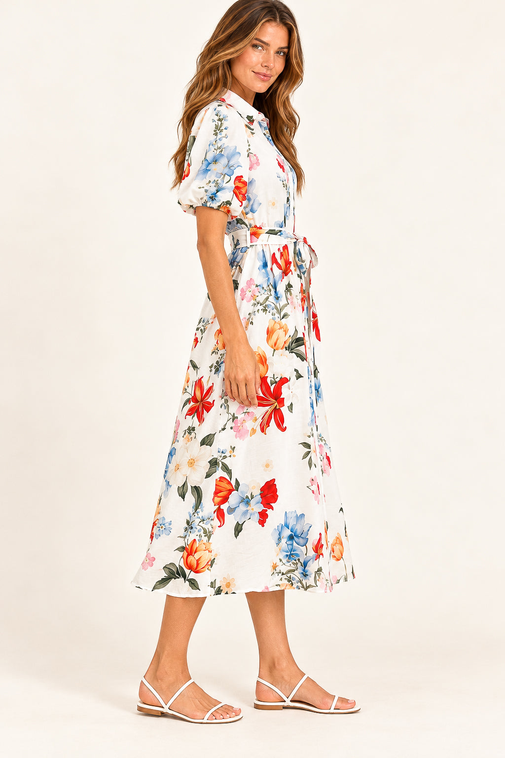 A woman modeling a white midi wrap dress with a vibrant multicolor floral print, featuring puff sleeves and a tie waist, paired with white strappy flat sandals against a plain off-white studio background.