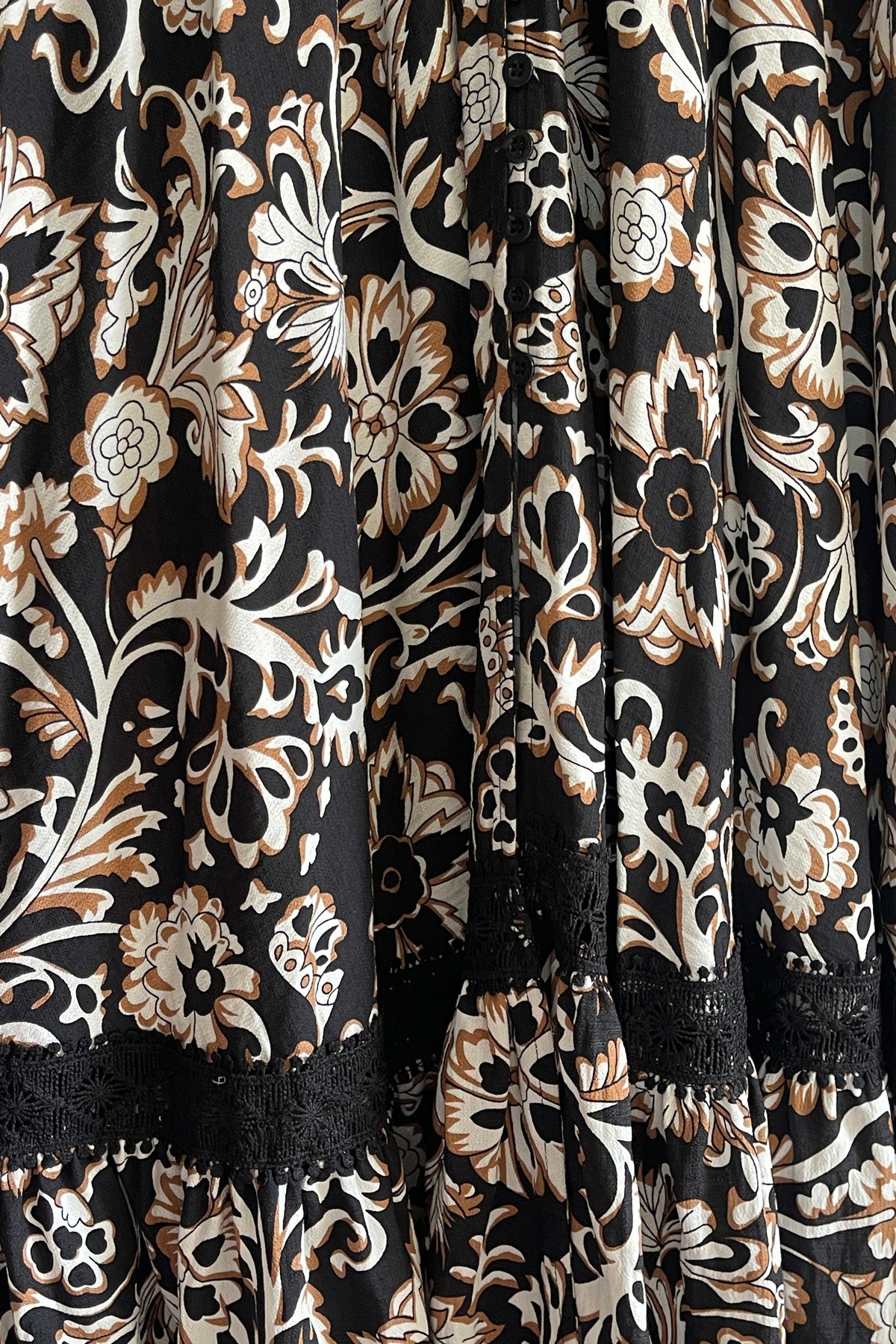 Close-up of a black tiered fabric with an intricate floral and scroll pattern in cream, beige, and brown, finished with black lace trim.