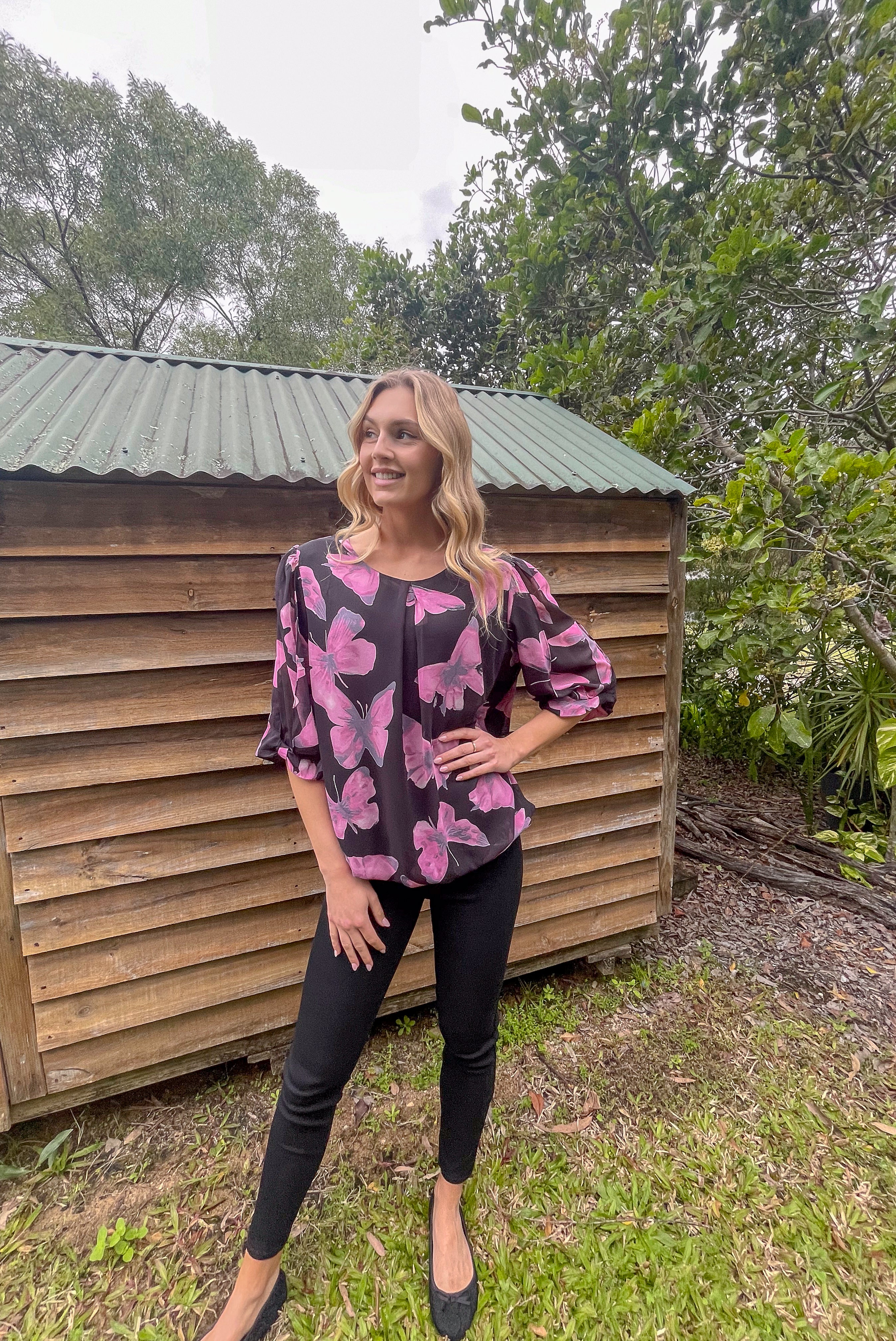 Woman wearing a black and pink floral blouse standing in front of a wooden shed with greenery around.