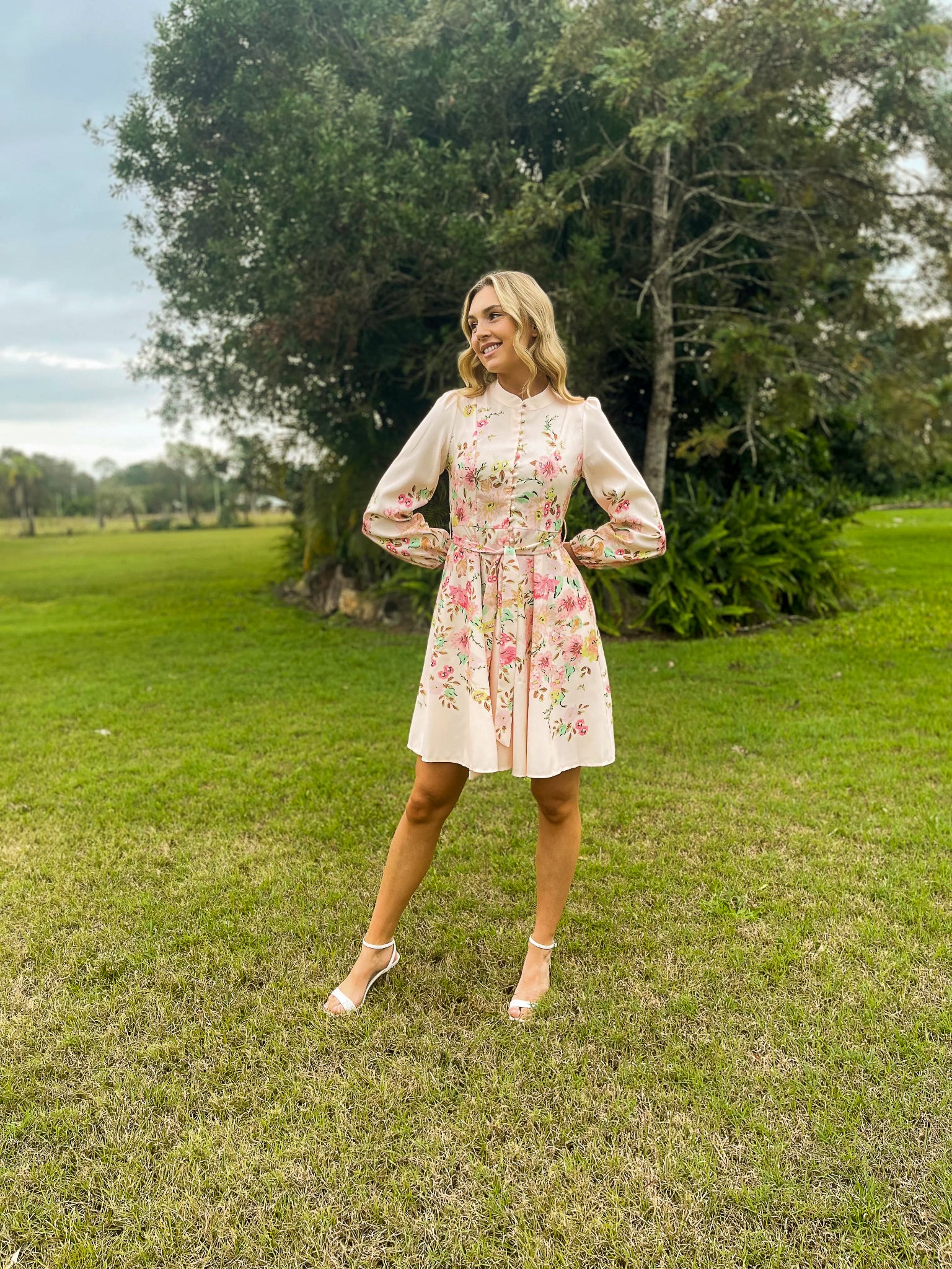 Woman in a floral dress standing in a grassy field with trees in the background