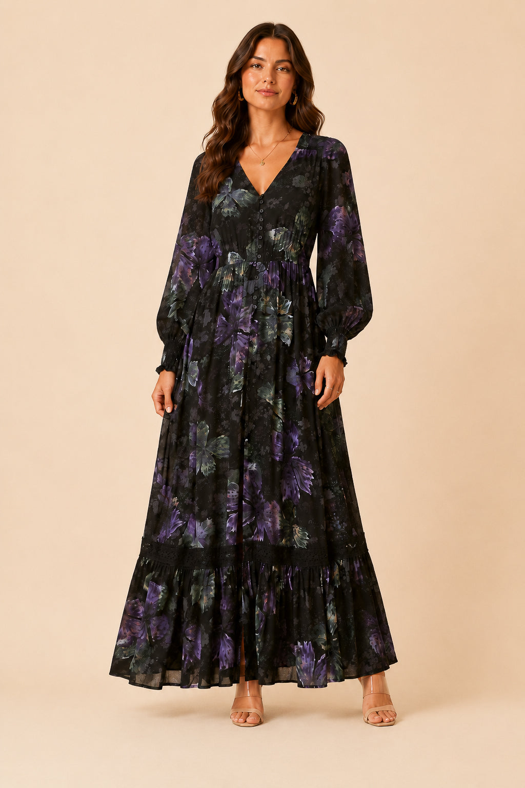 A woman modeling a long-sleeve maxi dress with a black base and a purple floral pattern, standing against a solid beige studio background.