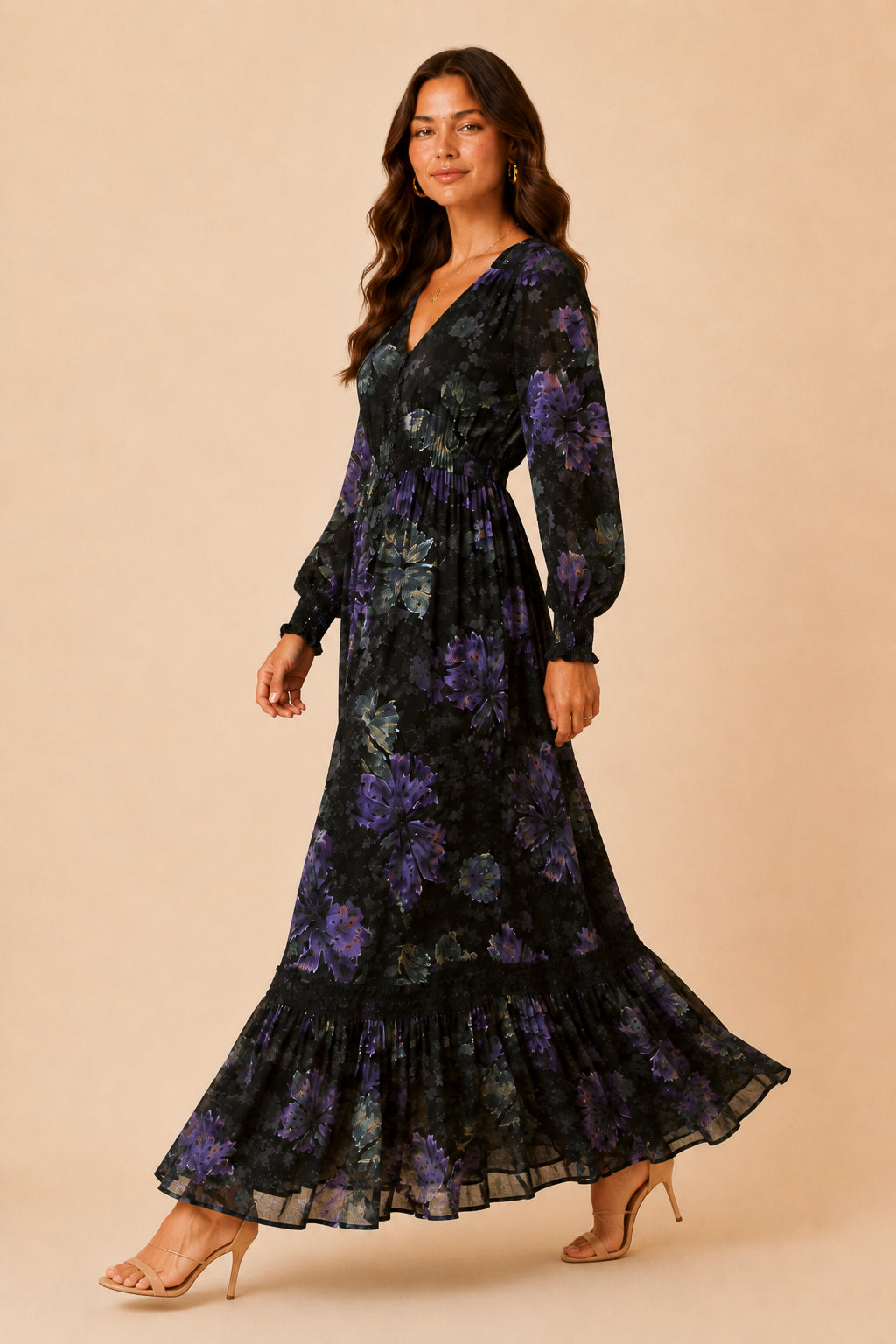 A woman modeling a long-sleeve maxi dress with a black base and purple floral pattern, standing against a solid beige studio background.
