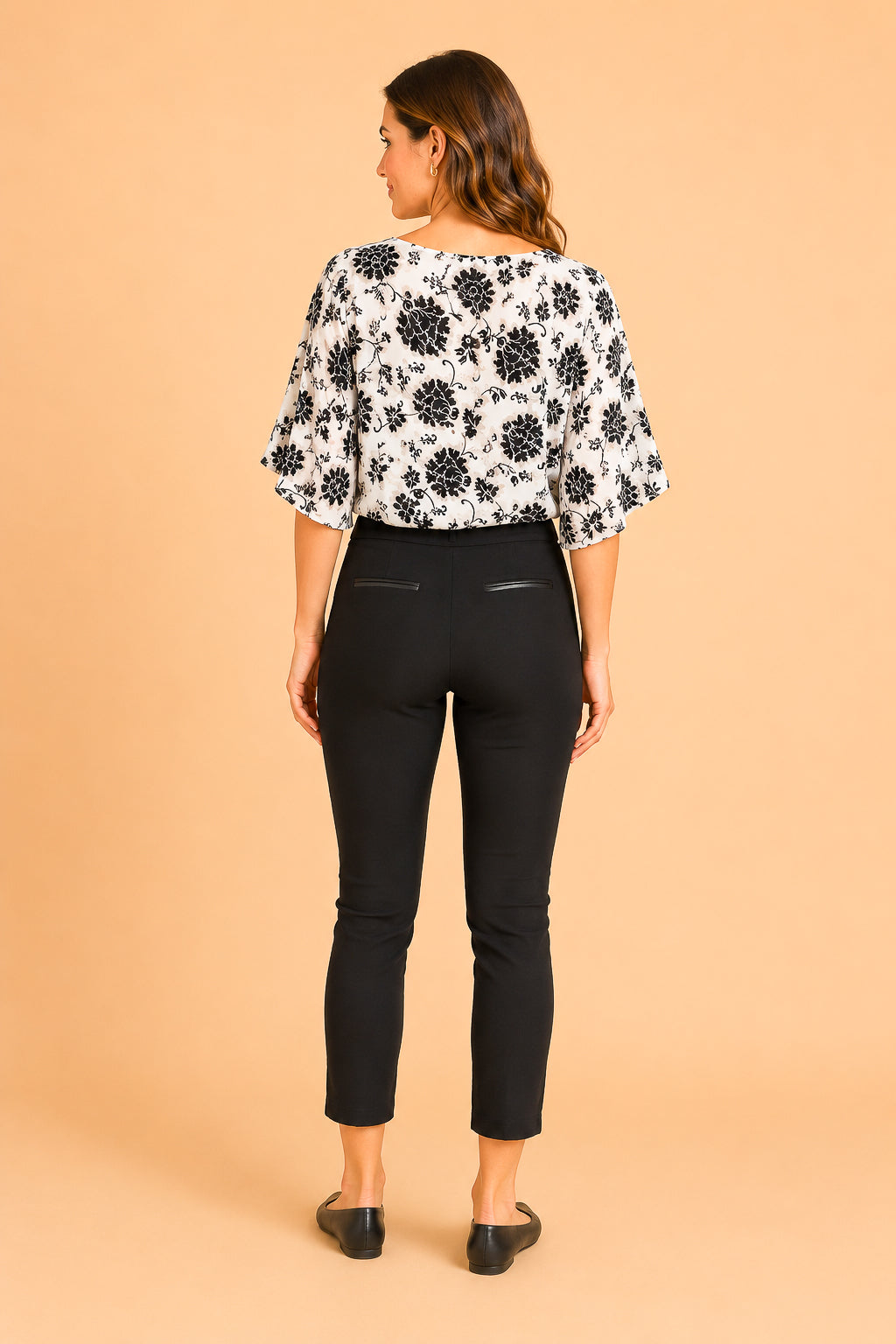 A woman shown from the back wearing a black and white floral patterned blouse with elbow-length puff sleeves and black cropped pants, standing against a solid peach studio background.