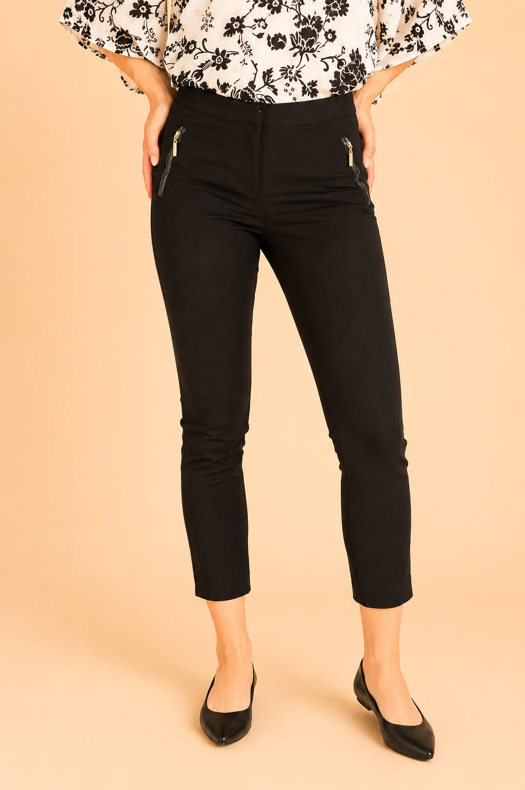 Black slim-fit ankle-length pants with zippered side pockets, shown on a model wearing a black-and-white floral blouse and black flats against a solid beige background.