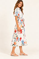 A woman modeling a white midi wrap dress with a vibrant multicolor floral print, featuring puff sleeves and a tie waist, paired with white strappy flat sandals against a plain off-white studio background.