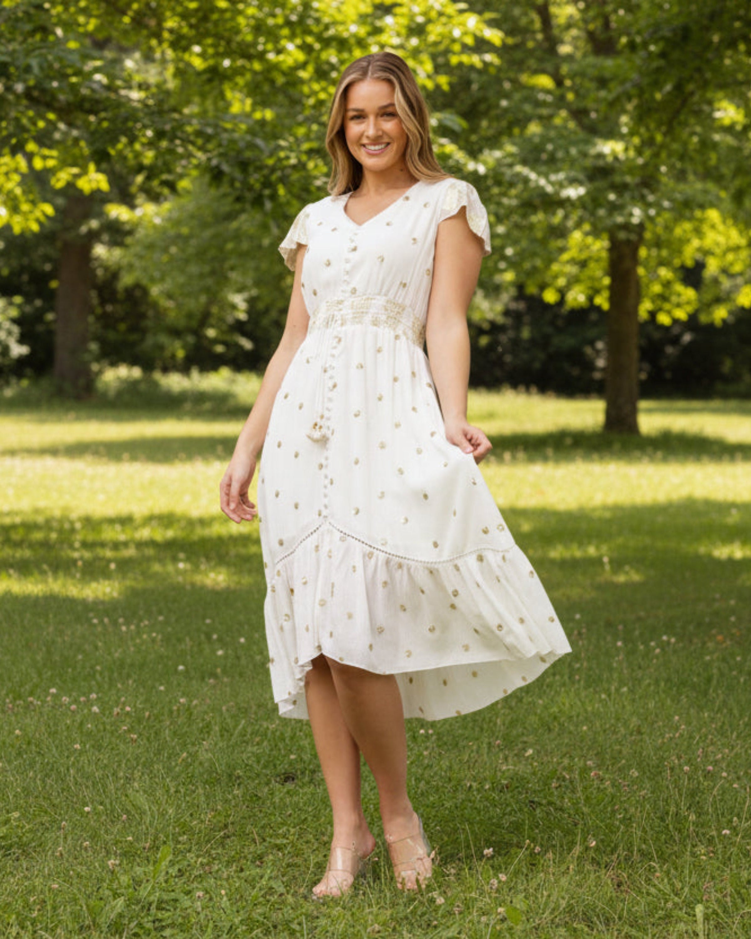 Woman in a white dress standing in a park with green grass and trees.