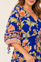 A close-up of a woman wearing a vibrant royal blue dress with a bold orange and yellow floral print, featuring puff sleeves with tie details and a buttoned V-neckline.
