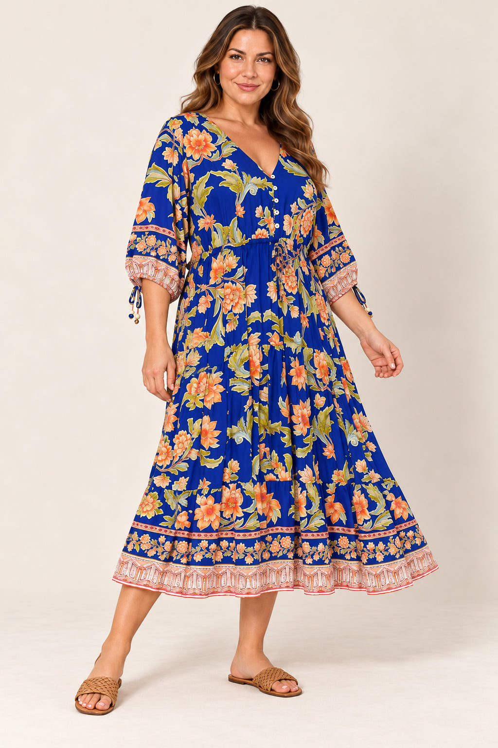 A woman modeling a royal blue midi dress with a vibrant orange, yellow, and green floral print, featuring three-quarter sleeves with tie details and a ruffled border, set against a plain beige studio background.