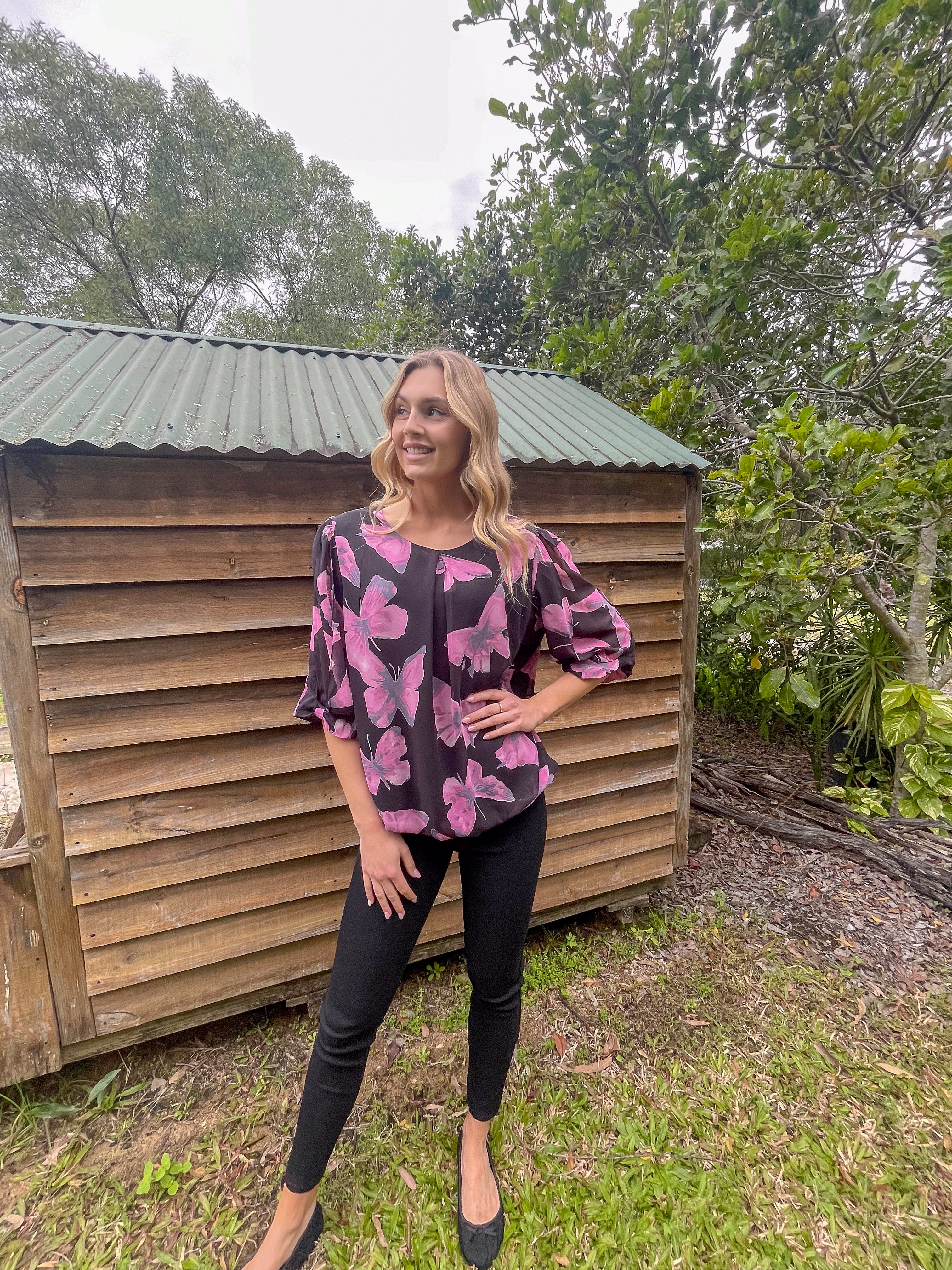 Woman wearing a black and pink floral blouse standing in front of a wooden shed with greenery around.