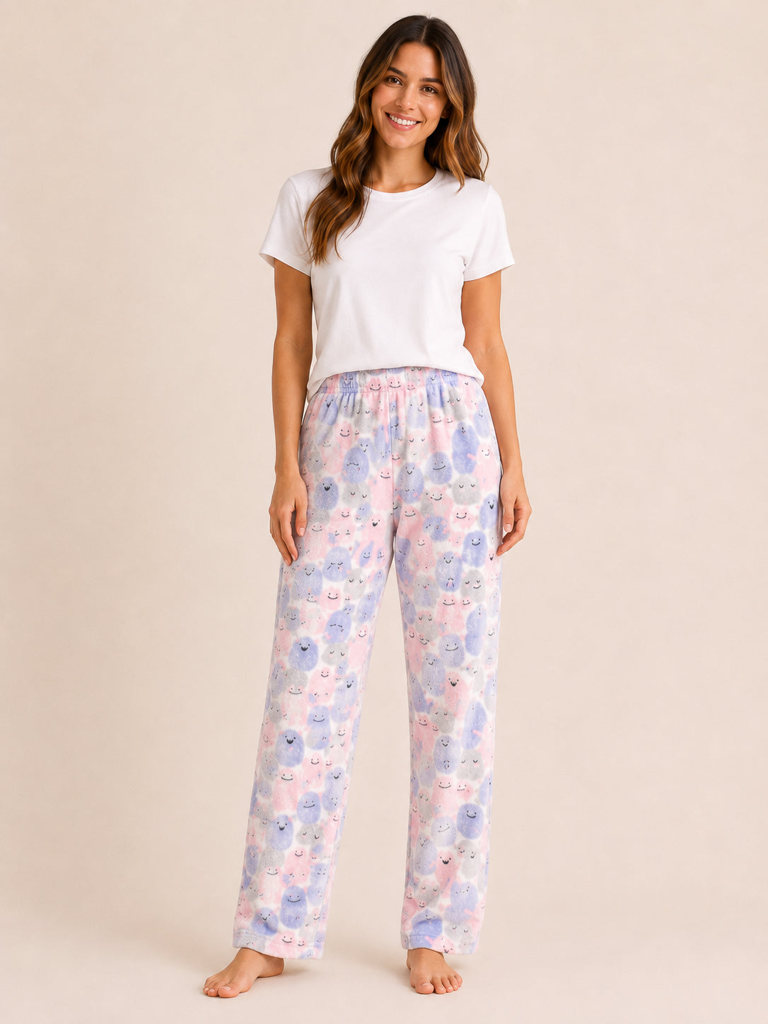 A woman modeling a pair of pajama pants with a pastel blue, pink, and purple cartoon animal print, paired with a plain white t-shirt, against a solid beige background.