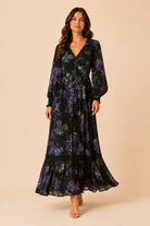 A woman modeling a long-sleeve maxi dress with a black base and purple floral pattern, standing against a solid beige studio background.
