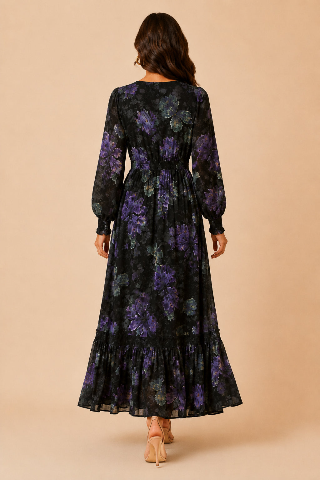 A woman viewed from behind wearing a long-sleeve black maxi dress with a purple floral pattern, standing against a solid beige background.