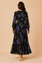 A woman viewed from behind wearing a long-sleeve black maxi dress with a purple floral pattern, standing against a solid beige background.