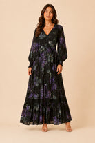 A woman modeling a long-sleeve maxi dress with a black base and a purple floral pattern, standing against a solid beige studio background.