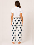 A woman viewed from behind wearing white wide-leg pajama pants with a navy blue star pattern, paired with a white short-sleeve t-shirt, against a plain beige studio background.