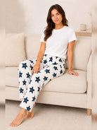 A woman sits on a light beige sofa wearing white pajama pants with a navy blue star pattern and a plain white t-shirt.