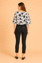 A woman shown from the back wearing a black and white floral patterned blouse with elbow-length puff sleeves and black cropped pants, standing against a solid peach studio background.